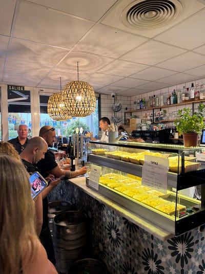 The photograph captures a scene in a tapas bar, possibly in Cambrils, Spain, judging from the Spanish text and the ambiance. The viewpoint is from within a crowd of patrons clustered at the bar. The counter is lined with metal beer kegs and a glass display case filled with tapa dishes. A menu card advertises tapas such as Ensalada de Pasta cubana and Torreznos at prices ranging from €4.50 to €10. A bartender stands behind the counter, visible above the crowd, attending to the customers. Patrons are engaged with the counter and their phones. Overhead, woven globe-shaped light fixtures illuminate the room. Through a window, the exterior is visible, with Cuban flags indicating the location. The interior is modern, with white tile walls and a geometric mosaic pattern on the walls behind the kegs. A circular ventilation outlet is set high in the upper left of the frame.