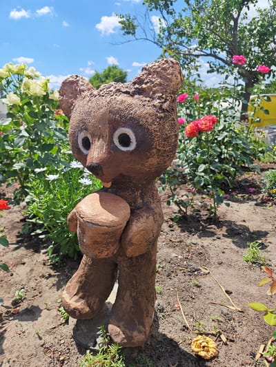 A close-up, low-angle outdoor photograph captures a decorative garden gnome resembling a bear, placed in a garden setting. The gnome is brown, with painted white eyes with black pupils, and is holding a brown cylindrical object. It stands on a bed of soil and small pebbles, with scattered dry leaves on the ground. Behind the gnome, a vibrant garden blooms with various flowers. To the left, white and yellow roses are visible, while to the right, clusters of pink and red roses are in full bloom. Green foliage and bushes are abundant, interspersed with small white flowers. In the background, a tree with green leaves stands tall against a bright blue sky dotted with fluffy white clouds. A hint of a yellow structure is visible in the far right background. The overall environment suggests a sunny day in a well-maintained garden. The gnome appears to be a static garden ornament, with no visible actions or interactions occurring. The location is identified as Krutinskiy, Russia.