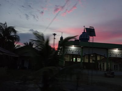 The image depicts a twilight scene in Blang Mee, Indonesia, likely during sunset given the vibrant pink and orange hues in the sky. The sky is filled with soft, scattered clouds, some tinged with pink, suggesting the lingering glow of the setting sun. A faint contrail stretches diagonally across the upper portion of the sky.
Dominating the right side of the frame is a building, possibly a mosque, characterized by a distinctive dome topped with a crescent moon and star symbol. The building's facade is illuminated, revealing a series of decorative architectural elements. In front of the building, palm fronds from several trees are silhouetted against the colorful sky, creating a strong contrast.
To the left of the mosque, a tall, lattice-like structure resembling a tower or a mast stands prominently. Further to the left, a portion of another building, possibly a house, is visible, partially obscured by palm trees and the dim lighting. In the foreground, the scene is dark and somewhat blurry, suggesting motion, possibly from a vehicle. A small vehicle with a sidecar is parked in front of the mosque.
The overall mood of the image is serene and peaceful, amplified by the dramatic sky and the presence of what appears to be a religious building. The dim light indicates it is either dawn or dusk, with the colors of the sky strongly suggesting sunset. No text is clearly visible in the image.