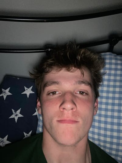A young man with brown hair and a green shirt is laying on a bed. He is looking directly at the camera with a neutral expression. His hair is messy, and there is a blue and white plaid pillow to the right of his face. There is also a pillow with white stars on a blue background to his left. The image is taken indoors, likely in a bedroom. The coordinates provided, 52.674, -2.466, are in Telford, United Kingdom.