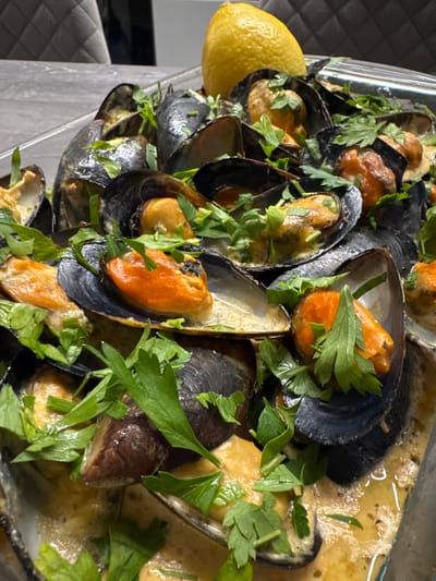 A close-up, overhead shot captures a dish of mussels bathed in a creamy sauce, garnished with fresh parsley. The mussels are dark-shelled and open, revealing their orange flesh. A whole lemon rests in the upper right corner of the frame, adding a touch of yellow to the scene. The dish appears to be served in a clear glass container, set against a blurred background that suggests an indoor dining setting, possibly a restaurant or home in Sustaš, Montenegro. The lighting is warm and natural, highlighting the textures and colors of the food. No people or other objects are clearly discernible.