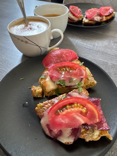 A close-up, eye-level shot shows a breakfast setting on a gray wooden table, located in Sustaš, Montenegro. In the foreground, a black plate holds two open-faced sandwiches topped with melted cheese, sliced tomatoes, and what appears to be thinly sliced cured meat, possibly prosciutto. Fresh green herbs are sprinkled on top. Behind these sandwiches, a white mug with a delicate black line design contains a frothy beverage, likely coffee or espresso, with a silver spoon resting inside. To the right of this mug, another white mug holds a lighter colored beverage. Further in the background, another black plate displays two more open-faced sandwiches, similar in presentation to those in the foreground, also adorned with sliced tomatoes and cheese. The lighting is natural and the focus is sharp on the foreground subjects, with the background elements slightly blurred.