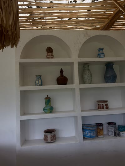 The image shows a series of built-in shelves in a white wall. The shelves contain a variety of decorative items, including vases and jars of different sizes, colors, and designs. In the top right shelf, there are also food jars. The left side of the image features a thatched or woven roof structure. The location is Tegal Suci, Indonesia.