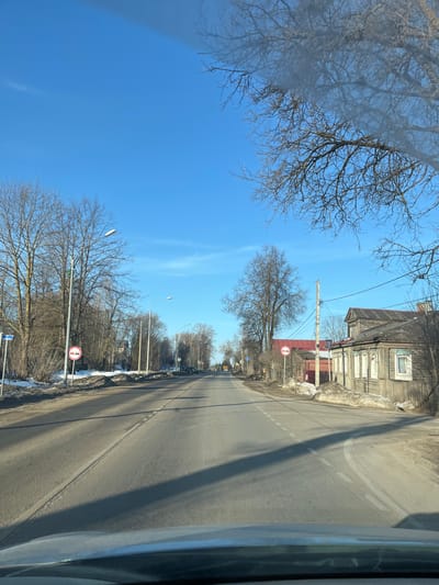 The image depicts a straight, asphalt road extending into the distance under a clear, bright blue sky in Maksimkovo, Russia. The perspective is from inside a vehicle, with the top of the dashboard visible in the bottom foreground. The two-lane road features a broken white centerline and is flanked by shoulders with visible remnants of dirty snow and bare, deciduous trees, suggesting a late winter or early spring setting. Long shadows are cast across the road surface from trees and structures, indicating direct sunlight.
On the left side of the road, multiple bare trees are visible, interspersed with streetlights. A blue street sign is partially visible near the foreground. A circular traffic sign with a red border, depicting two vehicles (one red, one black) side-by-side with a diagonal red line, indicating "No Overtaking," is positioned closer to the road. Further down the road, several parked vehicles are visible on the left shoulder.
On the right side of the road, similar bare trees line the shoulder, along with utility poles and overhead wires. Another "No Overtaking" traffic sign, identical in appearance to the one on the left, is visible. Adjacent to this sign stands a weathered two-story wooden house with multiple windows, some featuring decorative trim, presenting a traditional architectural style. Behind this house, a smaller, red-colored building is partially visible. No individuals or active interactions are visible within the scene.