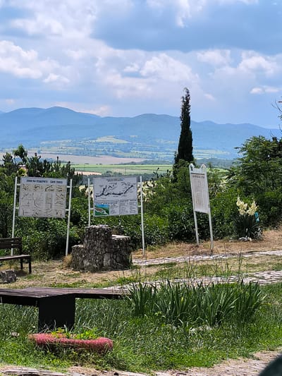 This image captures an outdoor informational area in the city of Raianovtsi, Bulgaria, overlooking a vast landscape. The scene is dominated by a clear, partly cloudy sky with numerous white cumulus clouds. In the background, multiple layers of mountains with a blue atmospheric haze extend across the horizon, with green and yellow agricultural fields visible in the valleys below.

The midground features dense green vegetation, including various bushes and trees. A tall, slender, dark green coniferous tree stands prominently in the center-right of the frame. Three white rectangular informational signs on metal stands are displayed:
1.  The leftmost sign is titled "ЗОНА НА ПЕЩЕРИТЕ" in Cyrillic and "CAVE ZONES" in English, featuring multiple diagrams and descriptive text below.
2.  The central sign presents a map-like diagram and additional descriptive text, predominantly in Cyrillic, likely pertaining to caves or the local area.
3.  The rightmost sign is primarily text-based, written in Cyrillic.

A low, rustic structure constructed from irregular stones is situated on the ground between the left and central informational signs. A dark wooden slatted bench with a metal frame is partially visible on the far left, and another similar bench is cut off at the bottom-left edge of the frame. A plant with multiple tall white flower spikes, resembling a Yucca, is visible near the rightmost sign.

The foreground consists of a mix of green grass and drier patches of earth. A light-colored paved pathway, possibly made of flagstones or similar material, winds through the grassy area. A red, tire-like object, potentially functioning as a planter, is visible in the grass at the very bottom-left, containing small green sprouts. No individuals are present in the scene.