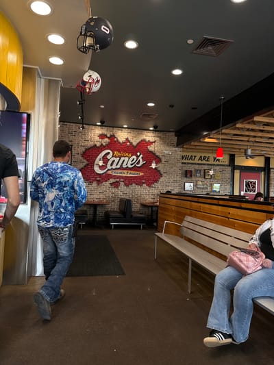 The image captures the interior of a Raising Cane's Chicken Fingers restaurant, likely in Mission, United States of America, illuminated by bright artificial lighting. The scene features multiple patrons and distinctive decor elements.

On the left side, a dark grey wall displays several recessed circular lights and square air vents. Two football helmets are prominently mounted: a black helmet with a white number "0" and a white helmet with red markings and face mask. The ceiling above has exposed wooden beams and metal structures.

The central and right sections of the restaurant showcase a brick wall. A large, red and white logo, depicting the "Raising Cane's CHICKEN FINGERS" name within a state-shaped outline, is a focal point. Above this logo, the phrase "WHAT ARE YA..." is partially visible in black letters. Several framed pictures and artworks are hung on the brick wall. A single red light fixture hangs from the ceiling in the mid-ground.

Several people are present. In the right foreground, a person is seated on a light-colored wooden bench, wearing light blue jeans and black sneakers with white stripes, and carrying a pink quilted handbag. Towards the center, a person stands facing away from the camera, dressed in a blue and white patterned long-sleeve shirt and jeans with embellished back pockets. This individual appears to be near a service counter or digital display, with another person's arms visible near the bottom edge of the frame. In the background, a person's head can be seen, possibly seated at another table or booth. The flooring throughout the restaurant is a dark, solid surface.