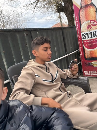 Here is a forensic description of the image:
A medium shot, rotated counterclockwise, shows a young man seated outdoors, possibly at a restaurant or cafe. He is wearing a light brown jacket with dark stripes on the sleeves and is holding a smartphone in his left hand. Another person, possibly a child, sits next to him, partially obscured by a dark blue puffy jacket. An advertisement for "Astika" beer is positioned above and behind the young man. The text on the advertisement includes "ASTIKA" and "FINE QUALITY LAGER" and appears to be written in Cyrillic script. A canopy-like structure is visible above, with trees and a cloudy sky in the background. The location may be in Zlatna Panega, Bulgaria, based on the Cyrillic text on the advertisement. The setting appears to be casual and relaxed, with the young man seemingly engaged with his phone.