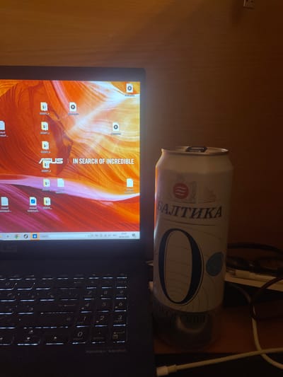 The image shows a laptop screen displaying a desktop with various icons, including a wallpaper with orange and pink abstract waves. Some of the icons are labeled in Russian, such as "202001_a" and "Новый текстовый". The ASUS logo and the slogan "IN SEARCH OF INCREDIBLE" are visible on the screen. To the right of the laptop, there is a can of "Baltika 0" non-alcoholic beer, which is white with blue and red accents. The can has the number "0" prominently displayed on it, along with text in Russian that appears to indicate it is non-alcoholic. Cables are visible behind the can. The laptop keyboard, labeled with white backlit keys, is in the foreground. The overall environment appears to be an indoor setting with a wooden desk. The presence of the laptop and beer suggests a personal workspace or leisure area. There is no information or visual evidence within the image that links the scene to the city of Laishevo, Russia.