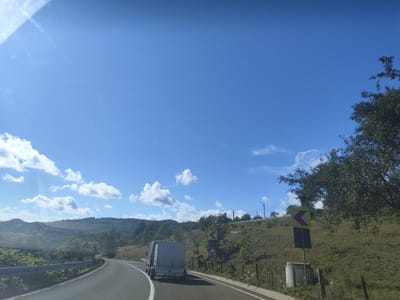 The image captures a view from a vehicle traveling on a paved road in a rural, hilly area. The road curves to the left in the foreground. A light-colored trailer is being towed by a vehicle in the same lane, slightly ahead. To the right of the road, a grassy embankment slopes upwards, dotted with trees and vegetation. Several road signs are visible on the embankment, including a yellow chevron sign indicating a curve to the left and a rectangular sign below it. A concrete structure, possibly a wellhead or culvert, is situated near the base of the embankment. The sky is bright blue with scattered white clouds. The background features rolling hills covered in trees and greenery. There is a guardrail along the left side of the road. The scene suggests a sunny day in a scenic, somewhat remote location, consistent with the landscape that might be found around Petrova, Romania.