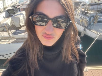 A female subject is depicted in a close-up selfie taken at a marina in La Grande-Motte, France. She has long, dark brown hair and wears black Ray-Ban sunglasses and a black turtleneck sweater. Her lips are pursed in a distinct "duck face" expression. The sunglasses reflect elements of the surrounding marina. In the background, two white sailboats are docked in dark water. The vessel to the left features a wooden deck and covered equipment, while the boat to the right shows visible rigging, ropes, and a fender. A wooden dock, marked with the numeral "3," is visible in the lower right foreground, indicating a berth number. The scene is under bright, direct sunlight.