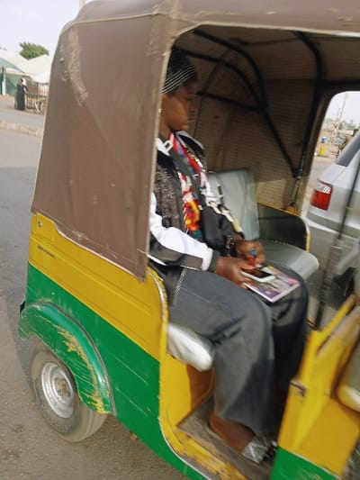 Inside a yellow and green auto rickshaw, a person reclines on the back seat, eyes closed, possibly asleep. A second person, wearing denim jeans and sandals, sits facing them, holding a tablet or book. The rickshaw's brown canopy is partially drawn. A tire is visible on the right side of the rickshaw. The background shows a blurry street scene with vehicles and buildings.