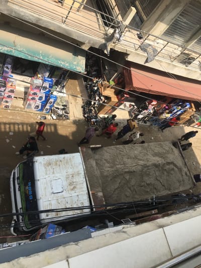 An overhead view depicts a bustling street scene in Ojo, Nigeria, under bright daylight casting strong shadows. A white and green SINOTRUK HOWO dump truck, loaded with a large quantity of light-colored sand or aggregate, occupies the center-left portion of the unpaved, potentially muddy street. Approximately 10-15 individuals, predominantly adult males, are visible on the street.
On the left side of the street, an open-front shop displays numerous stacked boxes, primarily containing electronics such as speakers and home theater systems. Near this shop, a person in a red shirt walks past the front of the truck. On the opposite side of the street, to the right, another commercial establishment features an extended reddish-brown awning and showcases boxed merchandise. Between these two main commercial areas, a vendor stall displays a large collection of shoes on the ground and rudimentary shelving.
Several individuals are gathered around the rear and passenger side of the dump truck, observing or potentially interacting with it. Other pedestrians are dispersed along the street, walking or standing. Overhead, a complex network of black electrical or utility wires crisscrosses the scene, connecting to the surrounding multi-story buildings. The buildings feature exposed concrete and metal railings. The street surface appears uneven and possibly wet or muddy.