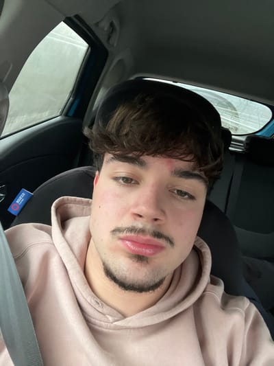 The image, oriented on its side, captures a young man, likely in his late teens or early twenties, taking a selfie or being photographed inside a car. He has dark, wavy hair styled to fall across his forehead, thick eyebrows, and brown eyes. A faint reddish mark, possibly an abrasion, is visible on his forehead. He sports a neatly trimmed dark mustache connected to a chin beard, resembling a Van Dyke style. His lips are full, and he looks directly at the camera with a calm, neutral expression. He is wearing a light beige or pale pink hooded sweatshirt.

The background shows the car's interior, including a dark headliner and part of a seat. Through a side window, an indistinct outdoor environment is visible, appearing somewhat blurred, which could indicate overcast weather or rain streaks on the glass. A blue tag or label with partially legible white text, appearing to say "SPARCO" and "CORSA RACING" or similar, is attached near the seatbelt buckle or on the seat itself. The photo was taken in Lamego, Portugal.