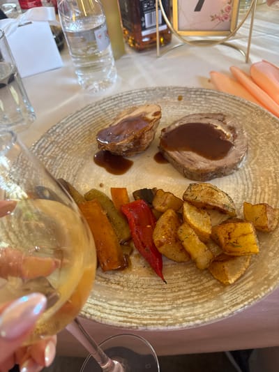 A close-up shot captures a delectable meal served on a rustic, patterned plate. The plate holds slices of roast meat drizzled with a rich brown sauce, accompanied by golden-brown roasted potatoes seasoned with dill. A medley of colorful roasted vegetables, including red bell peppers, zucchini, and carrots, adds visual appeal to the dish. In the foreground, a partially visible wine glass with a pearlescent sheen suggests a celebratory or dining occasion. The setting appears to be indoors, likely a restaurant or dining room, with a hint of a pale tablecloth and a blurred background. To the left, a clear plastic bottle of water and a decorative frame with the number "7" and floral accents are visible, hinting at a table setting for an event. A glimpse of peach-colored fabric in the upper left corner might indicate drapery or other table adornments. The lighting is soft, creating a warm and inviting atmosphere, possibly indicating an evening meal. The overall impression is one of a well-prepared and appetizing meal at a pleasant gathering.
