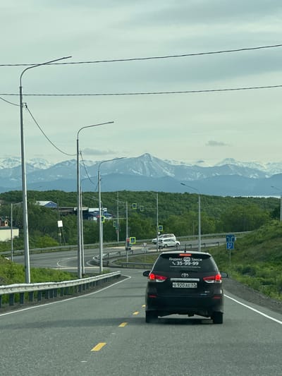 An overcast day in Krutoberegovy, Russia, features a black Toyota Wish station wagon with "Yandex Go Taxi" branding and a phone number on its rear window, driving on an asphalt road. The car's license plate reads "E 920 AE 41 RUS". The road curves to the left, bordered by a metal guardrail and green foliage on the right. In the distance, a dense green forest covers rolling hills, beyond which rise snow-capped mountains under a cloudy sky. Streetlights are visible along the roadside, as are road signs and traffic signals. A white SUV is parked on the left side of the road, further ahead.