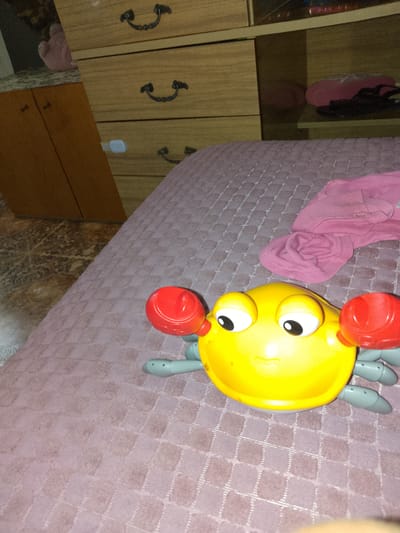 A yellow toy crab with red claws and large white eyes is positioned on a pink textured blanket. Behind the toy crab, a folded pink piece of fabric is visible. To the left, a wooden dresser with three drawers and ornate handles is partially visible, along with a wooden door. The scene appears to be indoors, likely a bedroom, in Manychskaya, Russia.