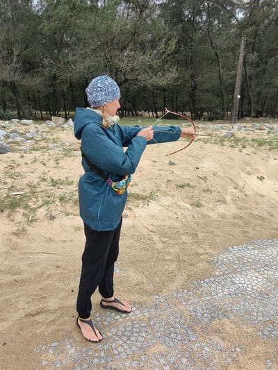 A female subject is depicted standing outdoors on sandy terrain in Thuận An Ward, Vietnam, aiming a small recurve bow. She is positioned with her body angled slightly to the right, wearing a light blue and white patterned head covering, a teal hooded jacket, a light-colored scarf, black trousers, and black minimalist thong sandals. A colorful patterned fanny pack with a black strap and buckle is secured around her lower back/hip. Her left arm is extended, holding a small reddish-brown recurve bow with a white string, while her right hand pulls back the string, an arrow visible nocked. The ground is composed of light brown sand with sparse green vegetation. Scattered light-colored rocks are visible to the subject's left. A patch of grey, patterned pavement, partially covered by sand, is present in the foreground to her right. The background features a dense line of tall, dark green trees and a dark utility pole with insulators and wires, under an overcast sky.