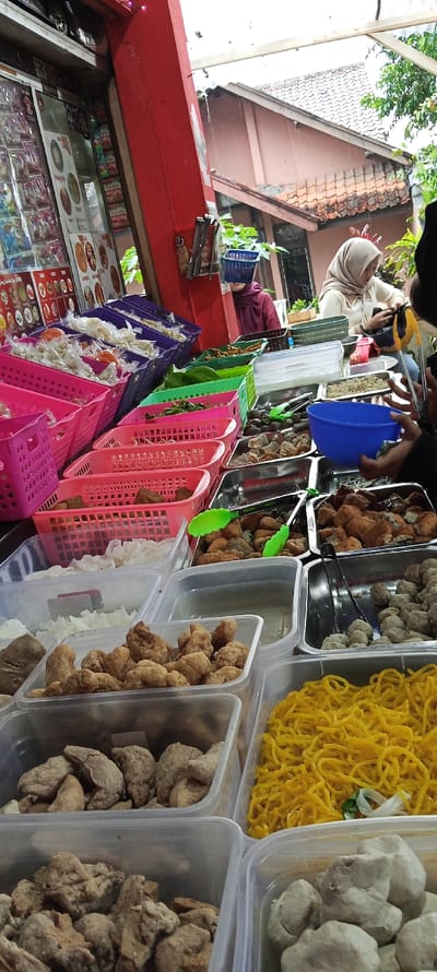 The scene is a street food stall in Cimaung, Indonesia. The stall is located on the street in front of a building with a red wall. There are several plastic containers filled with various types of food, including meatball-like items, deep-fried pieces of food, and noodles. One container has a blue lid and is filled with an unidentified substance.  A woman in a white headscarf and a blue shirt can be seen sitting to the right. A person wearing a black hijab can be seen from the back, their right hand is reaching for the food. The food is arranged on the table in front of them, and other customers can be seen in the background. There are many pink baskets full of food in the background. There are many different types of food in the stall, and the stall is busy with customers. The weather appears to be overcast.