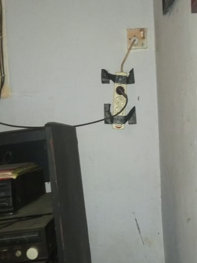 A vertical, off-white wall features a precarious electrical setup. At the top, a damaged wall outlet with a visible wire. Below, a power strip is secured to the wall with black tape. A plug is inserted into the top outlet of the strip. A black cable extends horizontally from the strip to the left, near a dark, dusty cabinet or shelving unit. The wall shows signs of wear and a streak of dirt.