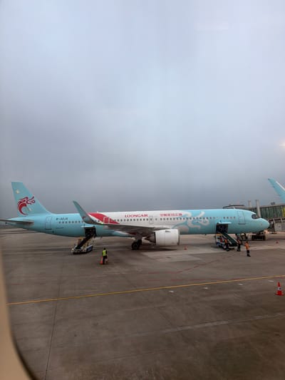 The image shows a Loong Air Airbus A320 aircraft on the tarmac at an airport, likely in Baiyun District, China, based on the airline's operating location. The aircraft has a light blue and white livery with a red and white design near the tail. Ground personnel in high-visibility vests are present near the aircraft, and a boarding stairs are connected to the front door. The ground is marked with yellow and red lines, and orange cones are visible. The sky is overcast.