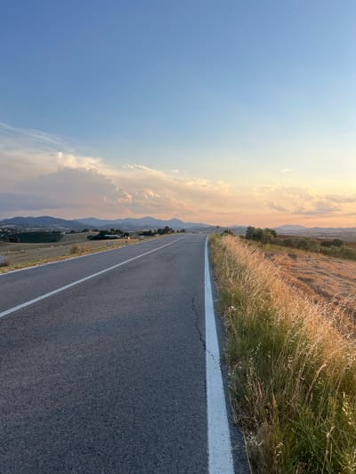 The image depicts a two-lane asphalt road with a white line marking the lane separation and a white line on the right shoulder, curving towards the right. The road is bordered by dry, golden-brown grass and some green foliage on the right side. In the background, rolling hills are visible, dotted with scattered buildings and olive trees. Further in the distance, a range of mountains forms the horizon. The sky is a gradient of blue, transitioning to pale yellow and orange near the horizon, with scattered clouds that are lit by the setting sun, indicating sunset or sunrise. The overall environment suggests a rural, possibly agricultural landscape. The scene does not contain any people or discernible actions. The location is identified as being in the vicinity of Ostra Vetere, Italy, a region known for its rolling hills and agricultural landscapes.