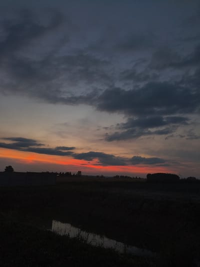 The image depicts a twilight scene over a rural landscape, likely in or around Galonki, Poland. The sky is dominated by dark, heavy clouds, with a band of vibrant orange and red light visible near the horizon, indicating the setting or rising sun. Below the sky, the land is silhouetted in deep shadow. In the foreground, there appears to be a ditch or small body of water reflecting the dim sky. Further back, a low, dark structure or wall runs horizontally across the frame, with trees and vegetation visible beyond it, also in silhouette. The overall impression is one of a somber, yet visually striking, natural environment at dusk. No people or specific actions are discernible.