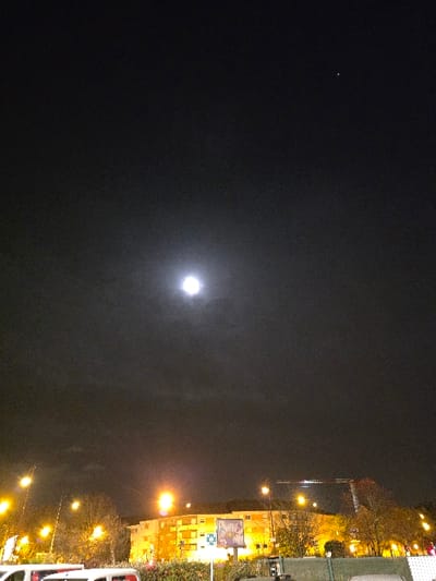 A night scene shows a cityscape against a dark sky. A bright moon is centered in the frame. Building lights and street lamps illuminate the right side of the image, creating a contrast with the dark sky. Trees are visible along the street. A few brighter building facades and signs add visual interest.
