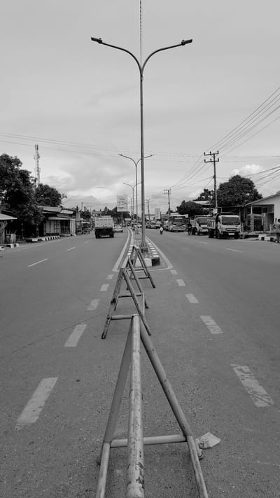 This is a black and white, low-angle, dutch tilt photograph of a street scene in Julikeude Dua, Indonesia. The image is oriented vertically, with the road stretching into the distance. The road itself is paved with visible asphalt texture and marked with dashed white lines. A metal barrier with a weathered, light-colored surface runs along the right side of the road.
The left side of the road is lined with trees and buildings, suggesting a built-up area. Several vehicles, including trucks and smaller cars, are visible, some of which appear to be parked or moving slowly. Overhead, a complex network of power lines crisscrosses the sky, supported by poles. A distinctive street lamp with a curved arm and a single fixture is prominent on the left. The sky is overcast with soft, diffused light, indicating it is likely daytime. There are no people clearly visible in the frame, but the presence of vehicles and buildings implies human activity.
Notable details include the texture of the road and the metal barrier, the dense arrangement of power lines, and the architectural style of the roadside buildings, which appear to be modest shops. The overall impression is of a typical, somewhat busy street in a tropical or subtropical region. The dutch tilt adds a sense of dynamism to the scene. No discernable text is visible in the image.