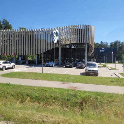 In the city of Berģi, Latvia, a contemporary building with a distinct curved wooden facade stands under a clear blue sky. The facade is composed of vertical wooden slats, creating a dynamic visual effect. Adjacent to this structure is a darker building with the "SKY" logo prominently displayed in white.
In the foreground, a parking lot is visible, filled with several vehicles including white SUVs, sedans, and a silver van. Some cars are parked under the covered section of the wooden building, which also features a "P" (parking) sign. A white SUV with red and white markings is parked on the left side of the lot, with the text "MUSTANG APCASTEF" visible on its side. A pedestrian crossing is marked with white stripes in the parking area.
The background features a line of evergreen trees, suggesting a natural or semi-rural setting. The ground in front of the parking lot is covered with green grass, and a paved walkway runs alongside it, with two circular manhole covers visible on the pavement. The overall scene depicts a modern commercial area set against a backdrop of nature.