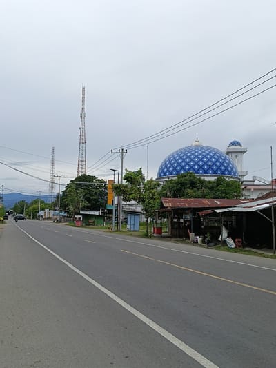 The image depicts a street scene in Paya Tringgadeng, Indonesia. A two-lane asphalt road runs diagonally from the lower left to the upper right of the frame, with a white solid line on the left edge and a broken yellow line in the center. On the right side of the road, there are several structures, including a prominent mosque with a large blue and white checkered dome and a smaller dome with a minaret. In front of the mosque, there are some low-lying structures with corrugated tin roofs, possibly market stalls or small shops. Various trees and foliage are present throughout the scene. In the background, there are communication towers, utility poles with wires, and a distant mountainous landscape under an overcast sky. A few vehicles, including cars and a motorcycle, are visible on the road, and some bags and containers are scattered near the roadside structures. The overall impression is one of a roadside view in a semi-urban or rural Indonesian setting.