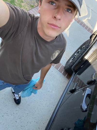 An outdoor, high-angle portrait captures a young male subject in a residential area of Rancho Palos Verdes, United States. The subject has light skin, blue eyes, and short brown hair, partially covered by a gray baseball cap worn backward. He is dressed in a dark brown short-sleeved t-shirt featuring a light-colored circular logo on the right chest, blue jeans, and black and white athletic shoes. A thin silver chain is visible around his neck, and a green-beaded bracelet is on his left wrist. He holds a light blue cloth in his left hand. To the right of the frame, a portion of a dark-colored vehicle, identifiable as a Jeep by its door hinges and handle, is visible, along with its front tire. The ground is a concrete driveway, bordered by bricks. Green grass is present in the upper left background. The scene is illuminated by strong natural light, casting long shadows indicative of either early morning or late afternoon. The subject is looking directly into the camera.
