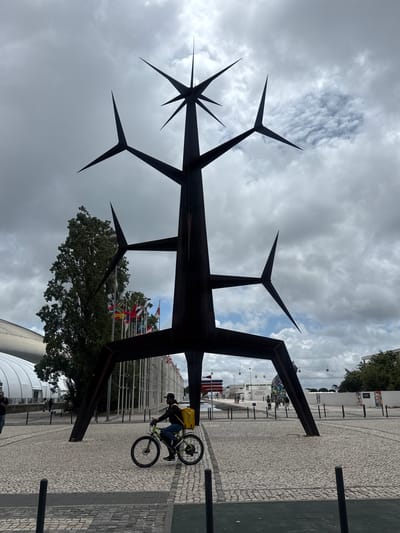 The image shows a large, abstract metal sculpture resembling a stylized tree with sharp, star-like branches, dominating the foreground. The sculpture is dark, likely made of steel, and is situated on a paved plaza. A person on a bicycle, wearing a black jacket and blue jeans, with a yellow delivery bag strapped to their back, is cycling across the plaza in front of the sculpture. The sky is overcast with dramatic clouds. In the background, there are several flags flying, a large green tree, and modern buildings, including a large white domed structure. The overall scene suggests an outdoor public space, possibly a park or exhibition area. This sculpture is known as the "Pavilhão de Portugal" and is located in Loures, Portugal.