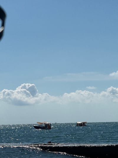 The image depicts a bright, sunny day at sea with a clear blue sky and scattered white clouds. The water is a deep blue and sparkles with sunlight, indicating choppy conditions. In the foreground, a dark, textured surface, possibly a breakwater or pier, juts out into the water. Further out on the water, two small boats are visible. The boat on the left has a light-colored canopy and appears to have people on board. The boat on the right is further in the distance and also has a canopy, with figures discernible within. The location is Adlerskiy, Russia.