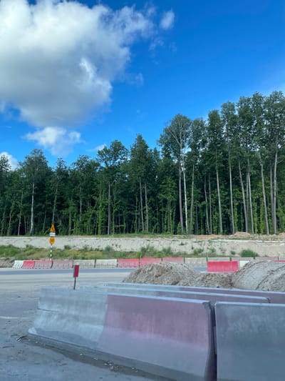 The image depicts a construction zone next to a forest, presumably in Novodrozhzhino, Russia. A line of tall, slender trees marks the forest edge against a vibrant blue sky with scattered white clouds. A roadway or path is bordered by alternating red and white concrete barriers. A yellow speed limit sign is visible. In the immediate foreground, large gray concrete slabs are stacked vertically.