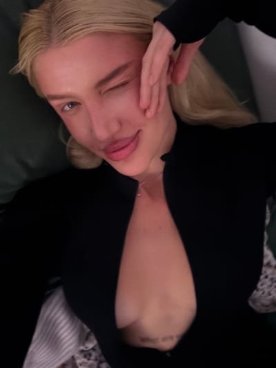An individual with blonde hair and fair complexion is depicted in a close-up, overhead shot, reclining in what appears to be an indoor setting. The person, likely female, is winking with their right eye while exhibiting a faint smile with plump lips. Their right hand is positioned with fingers splayed against the right side of their face, near the cheekbone. They are wearing a long-sleeved black garment featuring a deep V-neckline, which exposes a portion of their sternum and upper chest. A thin, light-colored necklace is visible around the neck. On the lower sternum, a small, dark-inked tattoo with text is partially visible. The immediate background behind the individual's head consists of a dark green, solid-colored fabric. To the lower right of the frame, a lighter-colored fabric with a subtle pattern is present. The specific location within the city of Yurlovo, Russia, cannot be determined from the visual information provided in the image.