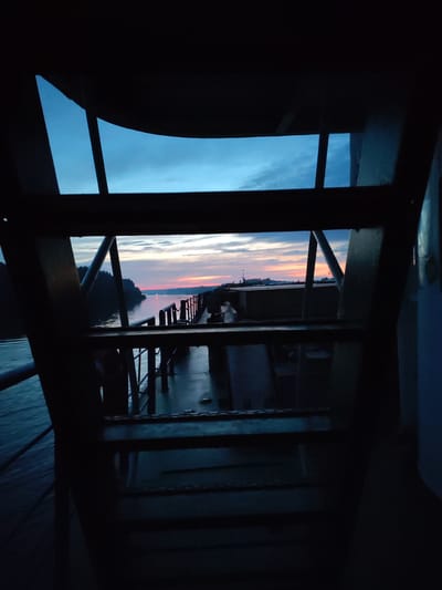 A wide-angle, low-light photograph taken from a ferry in Badozhskoi Pogost, Russia, captures a serene sunset over a body of water. The foreground is dominated by the dark silhouette of the ferry's interior, with metal railings and structural elements framing the view. The middle ground features the ferry deck, also in shadow, leading to a railing along the edge. Beyond the ferry, the water is calm, reflecting the soft colors of the twilight sky. A tree-lined shore is visible on the left, disappearing into the distance. The sky transitions from a deep blue at the top to soft hues of orange, pink, and yellow near the horizon, indicative of sunset. The overall scene evokes a sense of tranquility and the end of the day.
