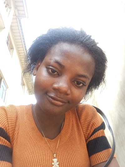 A close-up, upward-angled photograph depicts an individual, identified as a female with dark skin and dark, natural-textured hair, looking directly at the camera with a slight smile. She wears a ribbed, short-sleeved top in an orange-brown color with black horizontal stripes on the sleeves. A thin gold-colored chain necklace with a stone-encrusted teddy bear pendant is visible around her neck. Small stud earrings are present in both earlobes. A dark strap, possibly from a bag, rests over her right shoulder. The background is overexposed and largely out of focus, revealing architectural elements of a light-colored building with windows and a roofline to the left, and a plain light-colored wall to the right. The bright lighting suggests an outdoor daytime setting. The specific location within Agburu Uke, Nigeria, cannot be determined from the visual evidence provided in this image.