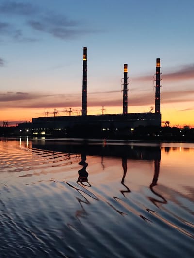 The image depicts a power plant with three tall smokestacks against a twilight sky at the Konakovo Power Station in Konakovo, Russia. The smokestacks are silhouetted against the colorful sky, with the topmost sections emitting a warm, orange glow. The power plant building is dark and indistinct in the mid-ground. In the foreground, the rippling surface of a body of water reflects the sky and the smokestacks, creating distorted, elongated shapes. The water appears dark and still, with subtle waves disturbing the reflections. The overall scene suggests the transition from day to night, with artificial lighting from the power plant contrasting with the natural colors of the sky.