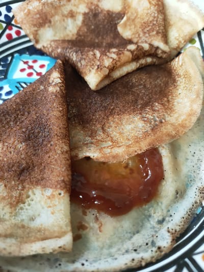 The image shows a close-up of crepes served on a colorful plate. The crepes are folded and appear to be filled with a bright orange jam or fruit preserve. The crepes have a slightly uneven, golden-brown color, indicating they have been cooked to a light crisp. The plate itself has a geometric pattern in blue, white, red, and green. The setting appears to be a casual meal, possibly breakfast or dessert, within a home or a cafe. The focus is on the texture and presentation of the food. There are no people visible in the image, and the lighting suggests an indoor setting. The image does not contain any visible text. The location context of Perpignan, France, might suggest a regional specialty, but this is not explicitly evident from the visual information alone.