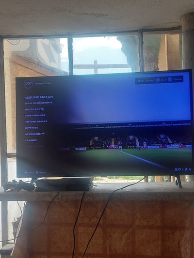 The image shows a television screen displaying a soccer game menu. The menu options include "Resume Match," "Team Management," "Match Facts," "Performance," "Instant Replay," "Settings," "Accessibility," "Trainer," and "Jump to Sim." The score is Burnley 0-2 Chelsea, with the time at 37:46. Advertisements for Burnley FC can be seen at the bottom of the screen. The television is located in a room with a window and patterned wall tiles. The location is Novo Zhelezare, Bulgaria. The window reveals a view of nearby buildings and the sky. A game controller is visible below the television.