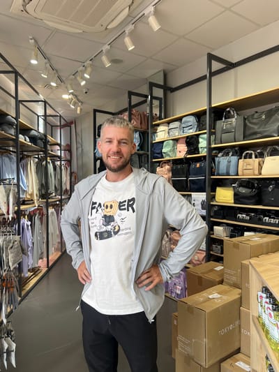 An indoor retail store in Thành phố Kon Tum, Vietnam, is depicted with a smiling adult male standing centrally. The man has short, light-colored hair and a beard, wearing a light grey zippered hoodie over a white t-shirt featuring a graphic of a cartoon character on a skateboard with the text "FASTER" and "TOKYOLIFE boarding teenagers", paired with dark trousers. He stands with his hands on his hips, facing the camera.
The store's interior features a white drop-tile ceiling with multiple track lights and a rectangular air conditioning unit. Shelving units with black metal frames and light-colored shelves line both sides of the aisle. The left side displays various articles of clothing on hangers, primarily in light colors, and some smaller textile items folded on lower racks. The right side features multiple shelves stocked with backpacks and handbags of different sizes and colors, including pink, blue, green, beige, black, and yellow. Below these are smaller accessories and several stacked brown cardboard boxes, some labeled "TOKYOLIFE". A mirror is partially visible on the right wall, and a small security camera is mounted on the ceiling track. The flooring appears dark and smooth.