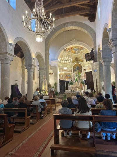 The image captures the interior of a church filled with congregants seated in pews. The church features a high, arched ceiling adorned with frescoes and elaborate chandeliers. Stone columns support the arches, creating a series of bays. The altar area is elevated and decorated with flowers and candles. People are sitting quietly in the pews, facing the altar, engaged in prayer or religious service. A red carpet runs down the central aisle. The setting is Rina, Italy, indicated by the architecture and cultural context.