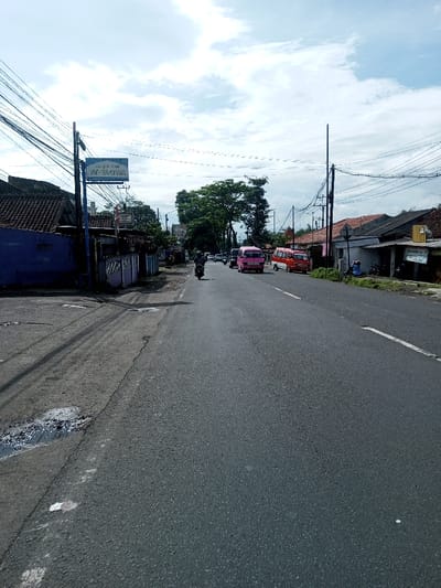 The image depicts a daytime street scene in Domphoog_Dungkarchhoeling, Bhutan, under a sky with scattered white clouds. An asphalt road, appearing to be two lanes wide, extends into the distance. On the immediate left, the road surface transitions to a rougher, possibly unpaved shoulder containing dark wet patches. A dark purple wall borders the left side of the road, behind which structures with reddish-brown tiled roofs are visible. A blue sign, partially obscured by overhead electrical wires, is mounted on a utility pole, displaying the text "SEJATI JAYA." Numerous black electrical or communication wires crisscross the frame horizontally and vertically, connected to multiple utility poles on both sides of the road.
A single individual on a black motorcycle, wearing a helmet, is operating the vehicle in the left lane, traveling away from the viewer. Their right arm is slightly raised. Further down the road in the right lane, a pink minivan is visible, followed by an orange minivan, both facing away from the camera and appearing to be either stationary or moving slowly. Beyond these vehicles, a large green tree stands prominently.
On the right side of the road, a series of single-story buildings with light-colored facades and reddish-brown tiled roofs line the street. A yellow rectangular sign is affixed to one of these buildings. Two individuals are seated on the ground directly in front of these buildings on the right side of the road. A continuous solid white line marks the right edge of the paved road surface.