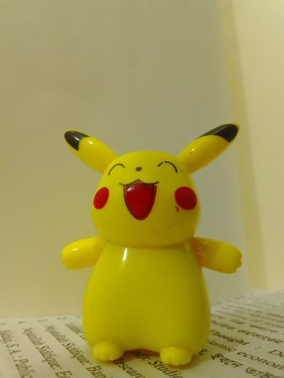 The image shows a yellow Pikachu toy figurine with a red tongue and red cheeks. It is situated on a table with a white background. The table has a book with black text on white paper underneath the figurine. The toy is located in the city of Rajshahi, Bangladesh. The coordinates (latitude: 24.366, longitude: 88.645) suggest the toy is located in a residential area of the city, near a book store. The figurine appears to be smiling with its mouth open, showcasing its red tongue. Its ears are black on the outside and yellow on the inside. The toy has two arms that are outstretched to its sides and two legs that are bent at the knees. The background of the photo is a white wall. The image was likely taken indoors, possibly in a living room or a bedroom. The toy is made of plastic. The book underneath the toy is open, and it appears to be a book of scientific or technical research. The image provides no further information about the location or the person who took the picture.