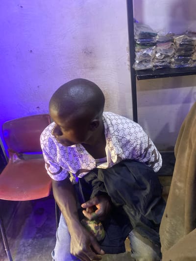 The image shows a person sitting on a red chair in what appears to be a dimly lit room. The person is wearing a white shirt with a repeating pattern of interlocking "G"s. A dark jacket is draped over their shoulder. The background includes a shelf filled with items, possibly phone cases or similar small products, wrapped in plastic. A purple light illuminates the left side of the scene, casting shadows. The person's head is bowed, and their arms are resting in their lap. The overall impression is one of a candid, perhaps casual moment captured in a commercial or retail setting. The presence of the items on the shelf suggests a store or market stall, while the lighting and seating hint at a less formal area, perhaps a back room or a waiting area. The time of day is not discernible, and no specific weather conditions are apparent. The scene is located in Barakin, Nigeria.