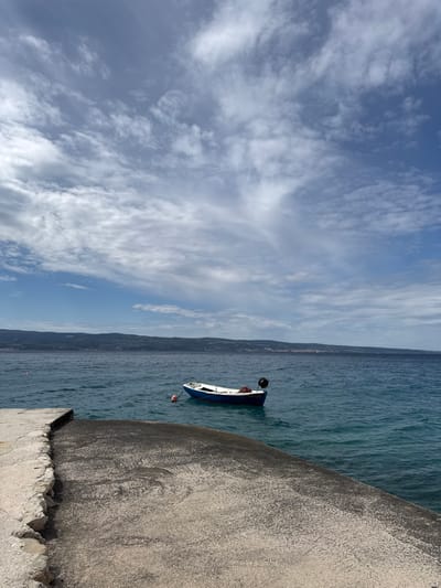 The image shows a single, small, blue and white boat moored in clear, blue water. It is docked at a small, concrete pier. The pier leads out into the water, and the boat is positioned a short distance away. There is a red buoy in the water, just in front of the boat. The background of the image is the Adriatic Sea, with a low, forested coastline in the distance. The sky above is a bright blue, with white, wispy clouds. The coordinates provided indicate that the location of the image is in Dugi Rat, Croatia. The scene is peaceful and serene.