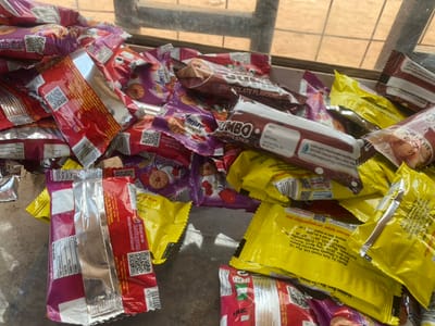 The scene, located in Okura, Nigeria, depicts a cluttered arrangement of numerous snack food packages and wrappers. The items are spread across a flat, light-colored surface, possibly concrete, under natural lighting. No individuals are present.
The objects primarily consist of assorted confectionery packaging. Prominently featured are:
* **Brown packages:** Identified as "JUMBO CHOCOLATE FLAVOURED STICK" or "CHOCOLATE STICK," manufactured by LIDA AGROPROCESSING NIGERIA LTD in Kano, Nigeria. Some display expiration dates and batch numbers.
* **Purple and Red/Pink packages:** Labeled "AMARE," these appear to contain biscuits or cookies, with imagery of circular biscuits and, on some, red berries/cherries. QR codes and social media logos (Facebook, Instagram) are visible on these wrappers.
* **Yellow packages:** Some are marked with "Noldon" and appear to be biscuits, manufactured by Yolo Foods Ltd. in Lagos, Nigeria.
* **Partially opened packages:** Several wrappers, particularly red ones, are crumpled or open, revealing silver foil interiors, indicating consumption or discard.
The environment suggests an indoor or sheltered outdoor setting, with a blurry reddish-brown background and dark structural elements visible in the upper portion, possibly part of a window or frame. The packages are in various states of integrity, from sealed to partially torn and crumpled.