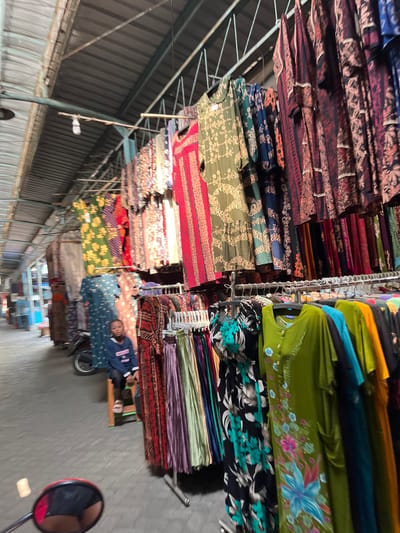 A young person sits on a small stool at a market stall in Juwet Kenongo, Indonesia. The stall displays a variety of colorful, patterned dresses hanging from racks, as well as other garments draped over a motorcycle. The market appears to be covered by a corrugated metal roof, with metal beams supporting the structure. The ground is paved with what looks like stone or concrete. The person, wearing a dark blue sweatshirt and light-colored sandals, looks towards the camera. The image is taken from a low angle, possibly from a motorcycle, as a red rearview mirror is visible in the foreground.