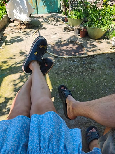 This image depicts a relaxed outdoor scene in Терновский округ, Ukraine, viewed from an elevated perspective looking downwards. Two individuals are present, represented by their legs and feet. In the foreground on the left, an individual with unclad legs is wearing a light blue dress with a white floral pattern and dark blue Crocs. The Crocs feature red and white striped soles and several decorative charms, including a Corgi, a smiley face, and other cartoon elements. This individual's legs are crossed at the ankles. To the right, another individual's leg and foot are visible, characterized by hairier skin. This person is wearing dark-colored, possibly rubber, slide sandals. A small portion of what appears to be denim shorts is visible on the right.
The ground consists of a mixture of concrete slabs and textured, darker earth with patches of moss. Strong shadows, indicative of bright sunlight, are cast across the ground from unseen foliage or structures. A coiled yellow garden hose lies across the ground from the upper left to the lower right.
In the mid-ground and background, various elements of a garden or backyard are visible. To the upper left, a white sheet or tarp is draped over a metallic object or frame. Behind this, a weathered, light green metal gate or wall is present. To the upper right, several green plants are contained within various pots and planters. Two large, light green, bowl-shaped planters, filled with dense green foliage, are prominent. Smaller brown and black pots are arranged beneath them. Behind these plants, a patterned fence or trellis with circular cutouts is discernible.
