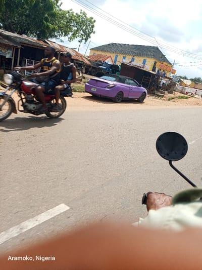 The image shows a scene from a street in Aramoko, Nigeria, captured from a motorcycle. The motorcycle is moving towards a purple convertible car parked on the right side of the road. Two men are riding the motorcycle, one driving and one seated behind him. The driver's hand can be seen on the handlebars and the passenger's hand is on the driver's shoulder. The motorcycle is moving forward on a paved road with a white painted stripe. On the right side of the road are several buildings, including a structure that is possibly a market or a shop. Trees can be seen in the background. The sky is clear with a few clouds.